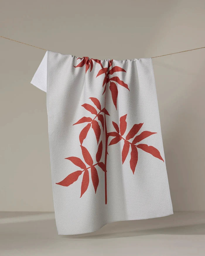 The "Rose" tea towel by PrintedPlant, featuring two large red leafy plant designs, hangs on a clothesline against a plain, light background.