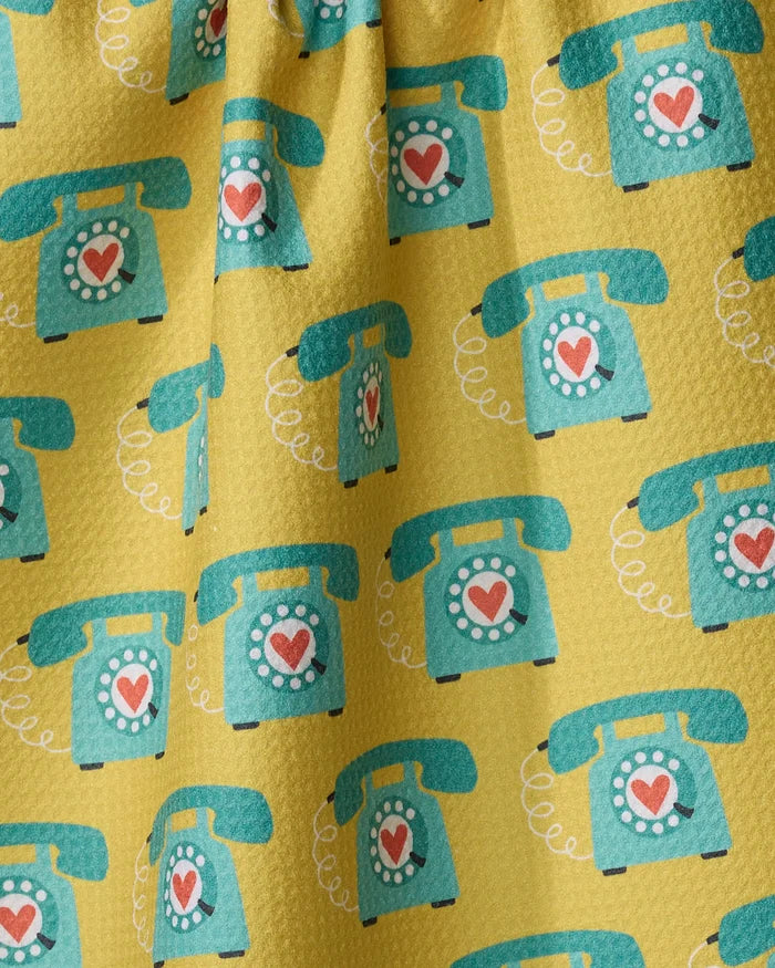 Megan Kampa's "Rotary Phones" tea towel features a yellow background with retro teal rotary dial telephones, each displaying a red heart and curling white cord, all crafted from recycled materials.