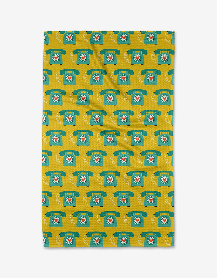 The Rotary Phones tea towel by Megan Kampa is rectangular, yellow, and made from recycled materials, featuring a green retro rotary telephone pattern with red centers. The towel is shown flat on a white background.
