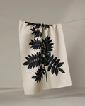 The Rowan tea towel by PrintedPlant, made from recycled materials, features a bold black botanical leaf print on beige and is shown hanging on a clothesline against a plain light background.