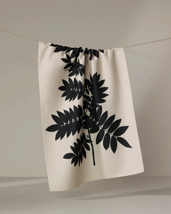 The Rowan tea towel by PrintedPlant, made from recycled materials, features a bold black botanical leaf print on beige and is shown hanging on a clothesline against a plain light background.