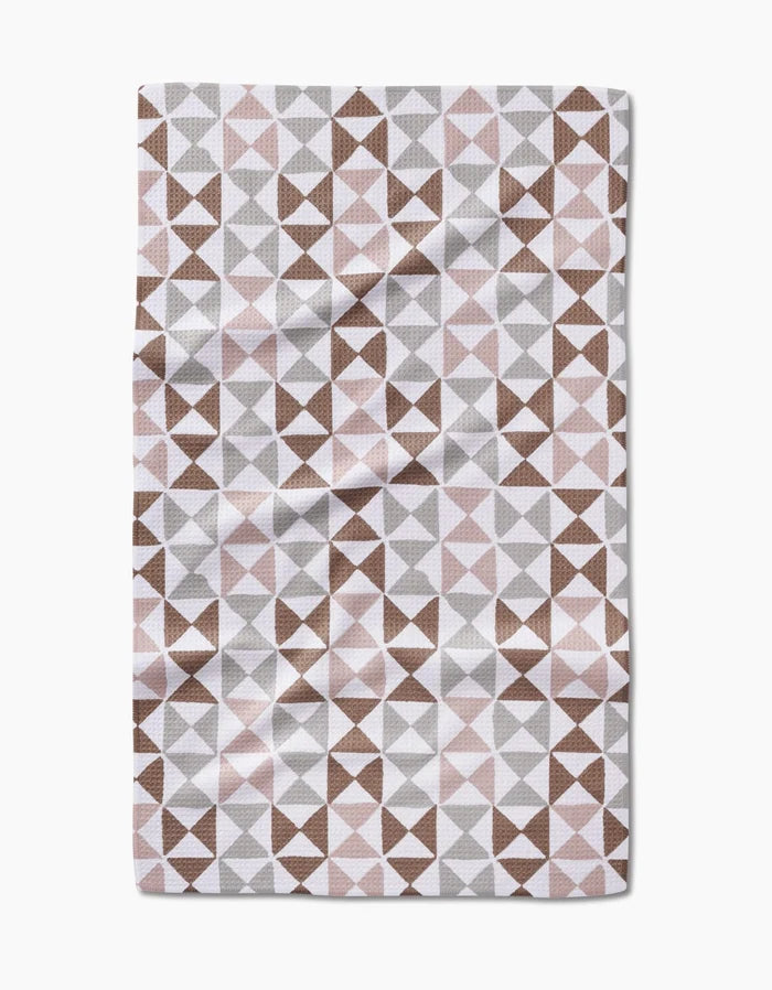 The Rubix tea towel by Studio Jeannot features a geometric triangle pattern in brown, gray, and light pink on Geoweave fabric made from recycled materials, shown with subtle creases against a white background.