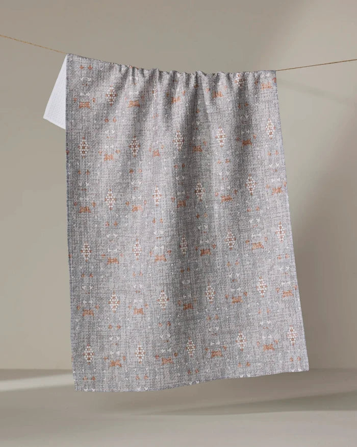 The Rulon Grey kitchen towel by Becki Owens, featuring a textured gray finish with white and rust geometric patterns, hangs neatly over a clothesline against a softly lit neutral background.