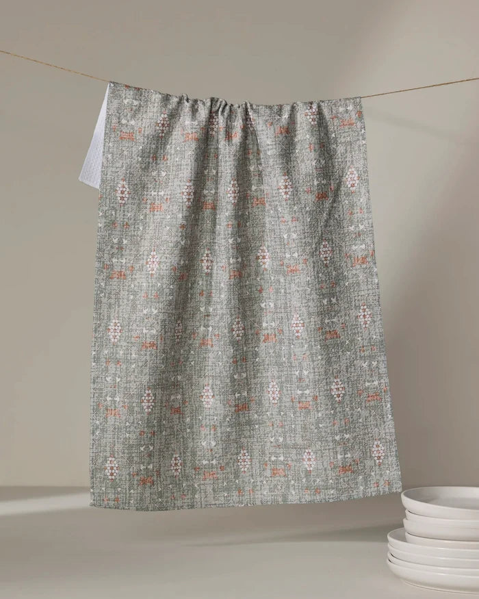 The Rulon Sage kitchen towel by Becki Owens, made from recycled materials and featuring a subtle geometric pattern in white and muted orange, hangs on a string beside a stack of white plates on a neutral surface.