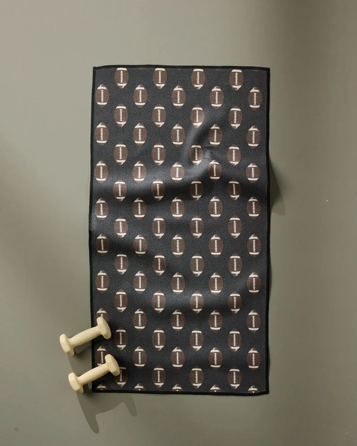 The Geometry Rush black fitness mat, made from recycled materials and featuring a repeating white “I” logo pattern, rests on a grey floor with two cream hand weights at the mat's bottom left corner.