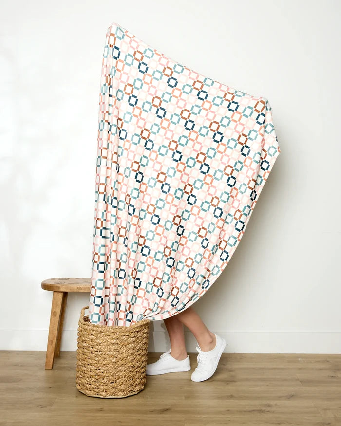 A person stands in a woven basket with only their legs visible, holding up the "Russet Crossroads" blanket by Charlie Rowan Designs, featuring geometric shapes in pink, blue, and black. A wooden stool sits nearby against a plain white wall.