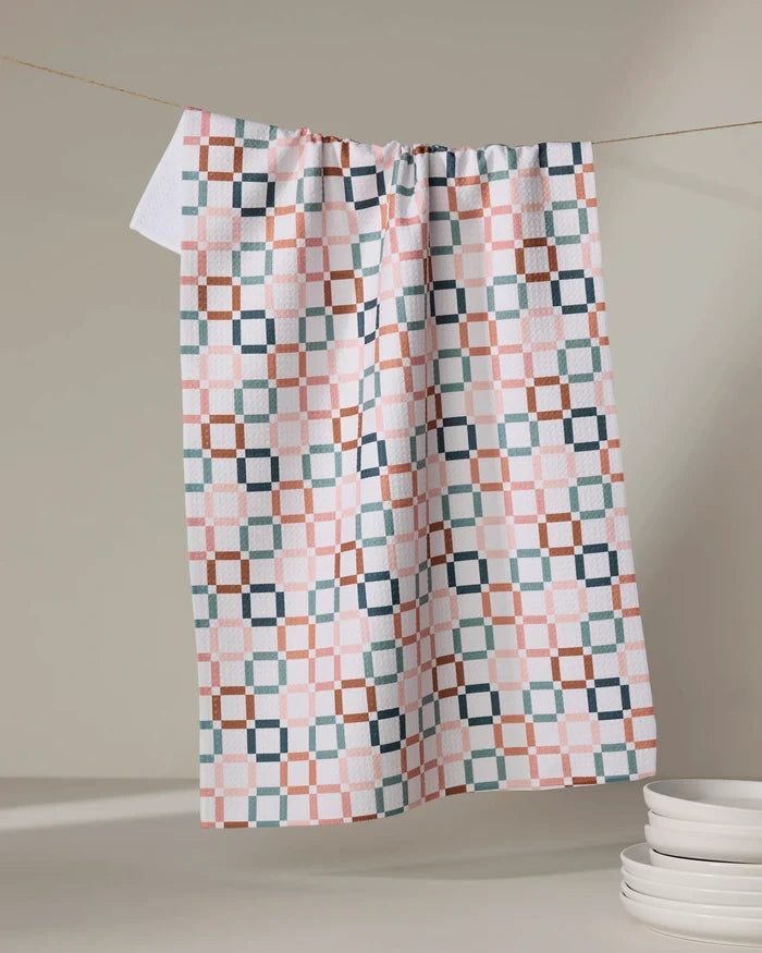 The Russet Crossroads tea towel by Charlie Rowan Designs, featuring a geometric pink, blue, and brown pattern, hangs on a line near stacked white plates, all set against a softly lit neutral backdrop.