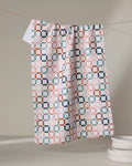 The Russet Crossroads tea towel by Charlie Rowan Designs, featuring a geometric pink, blue, and brown pattern, hangs on a line near stacked white plates, all set against a softly lit neutral backdrop.