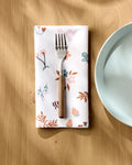 A fork rests on a folded Charlie Rowan Designs "Russet Meadow" reusable napkin with a floral pattern beside a light blue plate on a wooden table, as sunlight casts soft shadows across the scene.