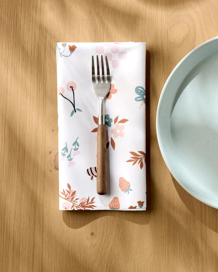 A fork rests on a folded Charlie Rowan Designs "Russet Meadow" reusable napkin with a floral pattern beside a light blue plate on a wooden table, as sunlight casts soft shadows across the scene.