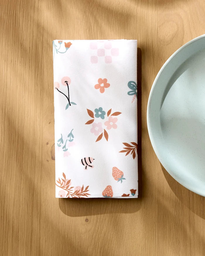 The Russet Meadow dinner napkin set by Charlie Rowan Designs, featuring colorful floral and berry patterns, is neatly folded beside a light blue plate on a wooden table with soft sunlight highlighting the charming scene.