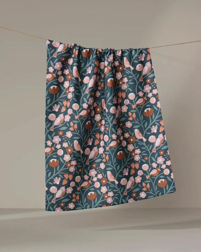 The Russet Park Lane tea towel by Charlie Rowan Designs, made from recycled materials, features pink and brown flowers and birds on dark green fabric and hangs on a string against a plain beige background.