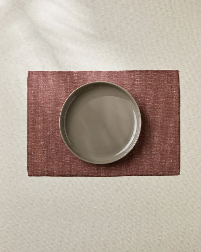 The Geometry Russet eco-friendly placemat in maroon, with a beige ceramic plate on top, rests on a light tablecloth, captured from above for a simple, minimalistic look.