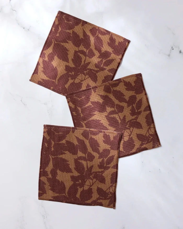 Three Rustle square cloth napkins by Geometry, featuring a brown and tan leaf pattern and made from recycled materials, are arranged overlapping on a white marble surface.