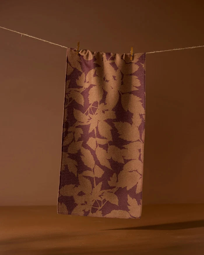 The Geometry Rustle recycled kitchen towel features a brown and tan leaf pattern, hanging on a string with two clothespins against a warm brown background.