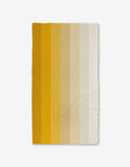 The GEOMETRY Saffron Levels is a rectangular blanket featuring vertical stripes in warm gradient tones from dark mustard yellow to off-white, crafted from recycled materials and shown on a white background.