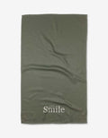 The Sage Smile tea towel by Kelsi Fullmer is green, textured, and made from recycled materials, featuring Smile printed in white near one edge and shown flat on a white background.