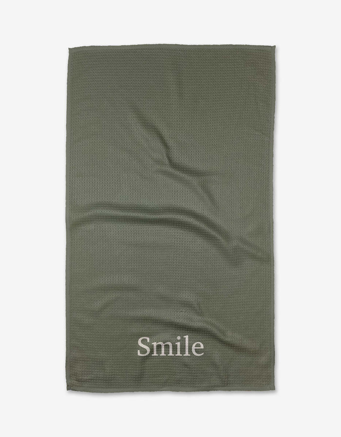 The Sage Smile tea towel by Kelsi Fullmer is green, textured, and made from recycled materials, featuring Smile printed in white near one edge and shown flat on a white background.