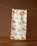 The Sagebrush Fox kitchen towel by Geometry, made from recycled materials, features orange and yellow foxes and green triangles in a repeating pattern. It hangs on a string with wooden clothespins against a brown background.