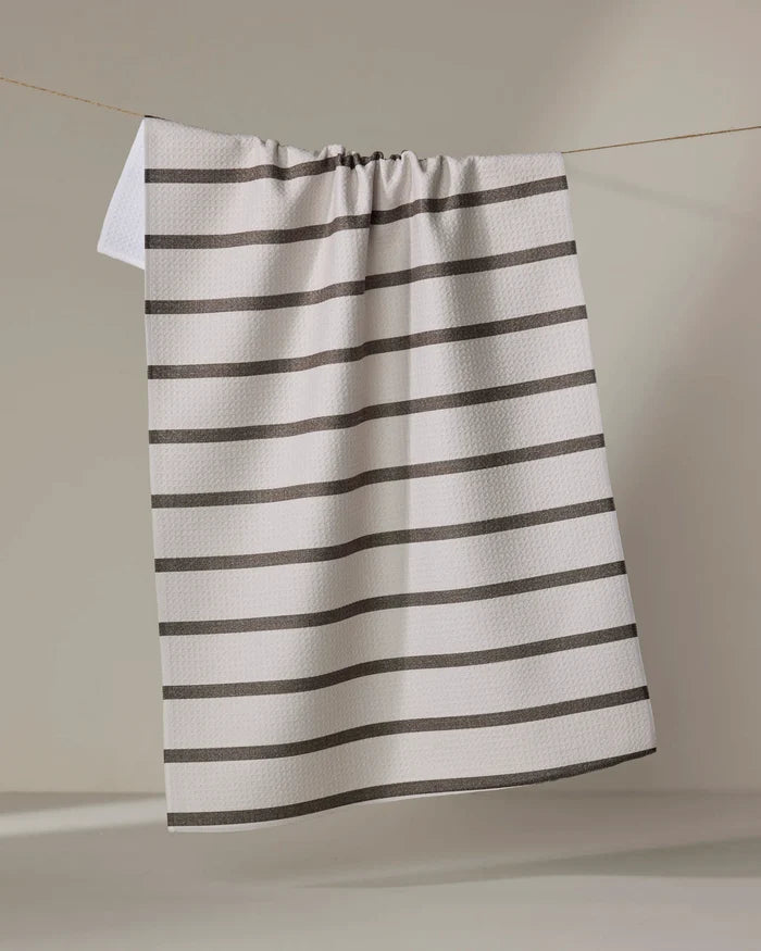The Jenny Keller Salt Air Stripe tea towel, featuring quick-drying white fabric with evenly spaced horizontal black stripes, hangs on a clothesline against a neutral background.