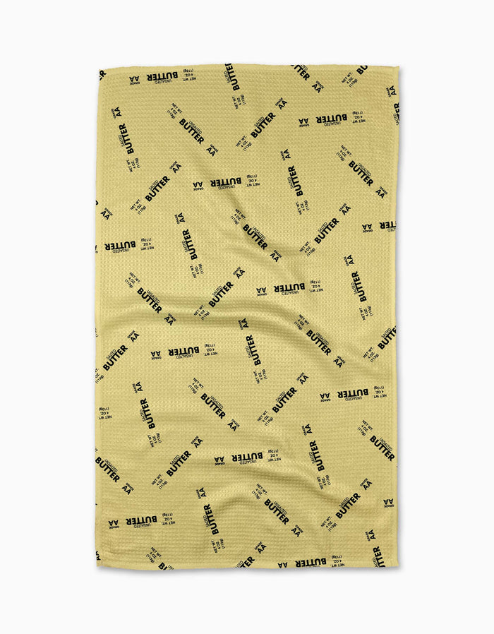 A yellow GEOMETRY Salted vs Unsalted Tea Towel with a waffled texture, scattered black “BUTTER” pattern, and small “XX” symbols. Made from recycled materials and shown laid flat with slight wrinkles.