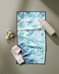 A flat lay of workout gear features Geometry's Saltwater Glow fitness towel with a blue water-like pattern, alongside a white water bottle, small dumbbells, and a white resistance band on a light grey surface.