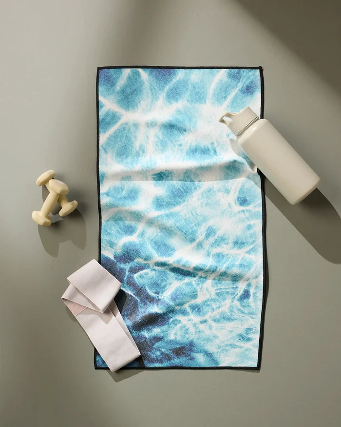 A flat lay of workout gear features Geometry's Saltwater Glow fitness towel with a blue water-like pattern, alongside a white water bottle, small dumbbells, and a white resistance band on a light grey surface.