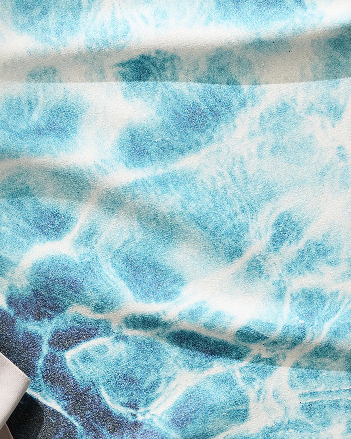 A close-up of Geometry’s Saltwater Glow fitness towel shows its blue and white wave pattern, ultra-absorbent soft texture, and a partially visible beige object in the lower left corner.