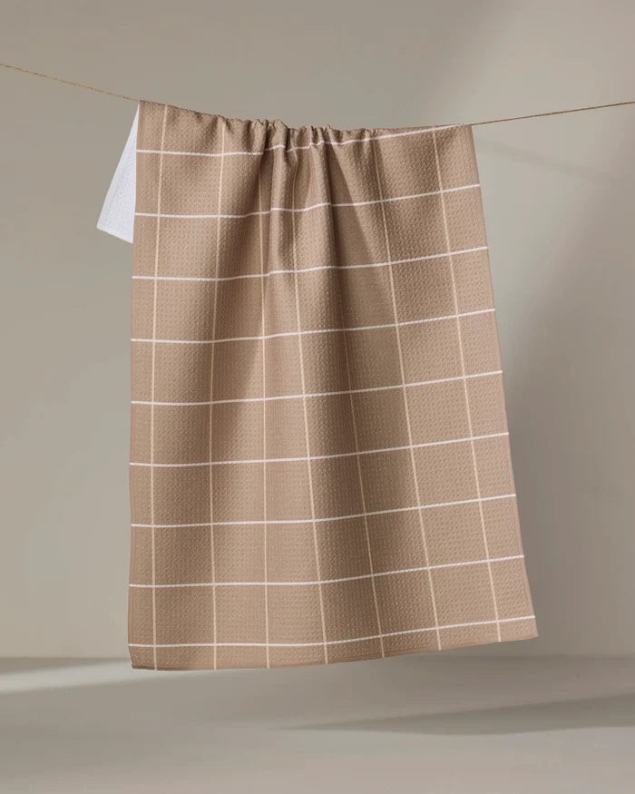 The "Sand Grid" recycled kitchen towel by Kelsi Fullmer, featuring a white grid pattern in beige, hangs on a line against a neutral background.