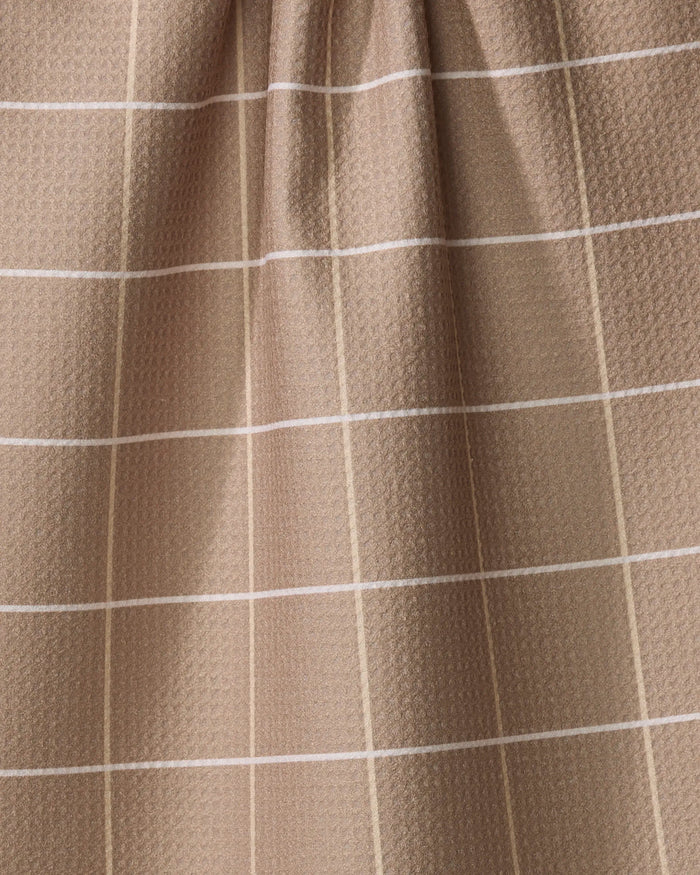 The Sand Grid kitchen towel by Kelsi Fullmer features a light brown recycled fabric with white and beige grid lines forming squares and a subtle textured pattern. The top is slightly gathered, creating gentle folds.