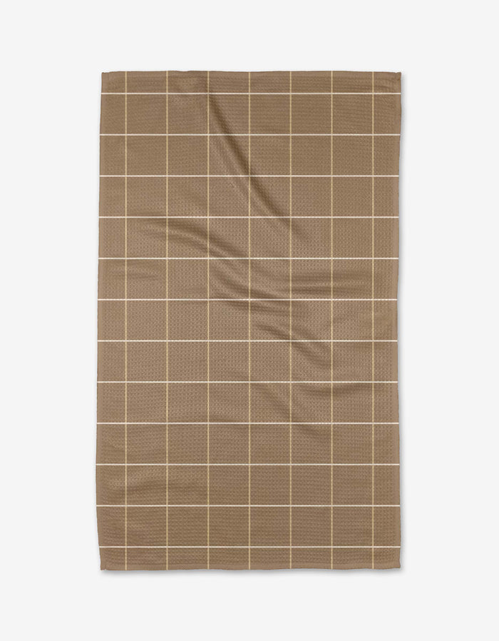 The Kelsi Fullmer Sand Grid Tea Towel is rectangular and brown, featuring a grid of thin beige lines on white Geoweave fabric, with subtle creases and folds visible.