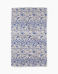 The Santorini Tile Tea Towel by Geometry is a rectangular beige towel with a blue nautical pattern of fish, seashells, starfish, and checkerboard designs. Made from recycled materials, it’s shown flat on a white background.