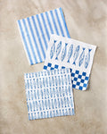 The Sardine Check Blue dishcloth set by Hufton Studio features three super-absorbent, recycled cloths with blue stripes, fish, and checkerboard designs, beautifully displayed on a light beige surface.