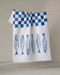 The Sardine Check Blue tea towel by Hufton Studio dries quickly and features a blue checkered top with six blue illustrated fish lined up vertically on the lower half.