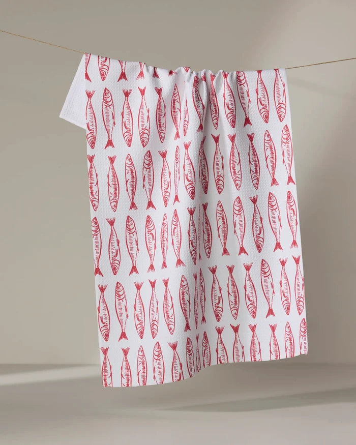 The "Sardine Stripes" tea towel by Hufton Studio features a repeating red fish pattern and is shown hanging on a clothesline against a neutral background.