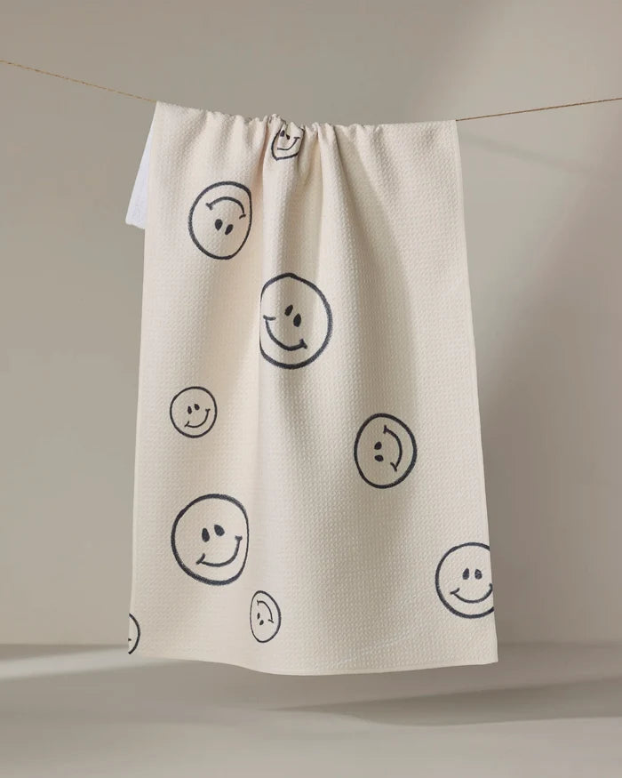 The Say Cheese tea towel by Kelsi Fullmer is cream-colored, made from recycled materials, and features black outlined smiley faces of various sizes on its textured, quick-drying surface.