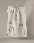 The Say Cheese tea towel by Kelsi Fullmer is cream-colored, made from recycled materials, and features black outlined smiley faces of various sizes on its textured, quick-drying surface.