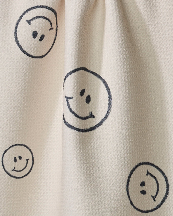 The Say Cheese tea towel by Kelsi Fullmer is cream-colored with a textured feel, decorated with scattered hand-drawn black smiley faces in various sizes, and made from recycled materials.
