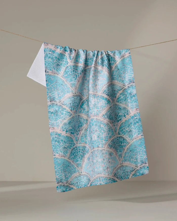 The Taryn Brennan "Scallop Hills" kitchen towel, featuring a blue and white scalloped pattern and made from recycled materials, hangs from a clothesline against a neutral background.