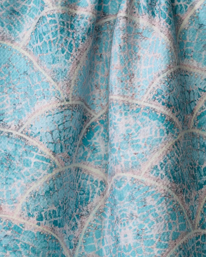 Close-up of the Scallop Hills tea towel by Taryn Brennan, showcasing its turquoise and white scale-like design with textured folds and soft fabric shadows.