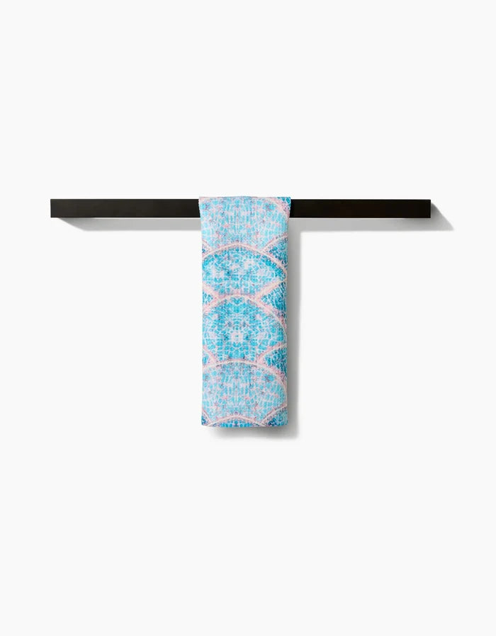 The Scallop Hills Bar Towel by Taryn Brennan, featuring a light blue and pink pattern and made from recycled materials, is draped over a sleek black towel bar against a plain white background.