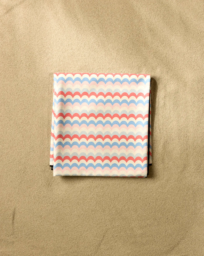 The Geometry Scallop Stripes beach blanket, featuring a pastel pink, blue, and white scallop pattern and made from folded micro-loop fabric, rests on smooth, light-colored sand.