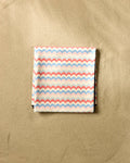 The Geometry Scallop Stripes beach blanket, featuring a pastel pink, blue, and white scallop pattern and made from folded micro-loop fabric, rests on smooth, light-colored sand.