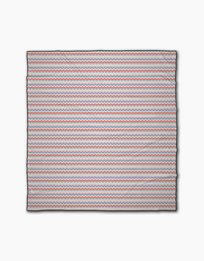 The Geometry Scallop Stripes is a square beach blanket made from recycled materials, featuring small red, blue, and white wavy stripes on a light background.