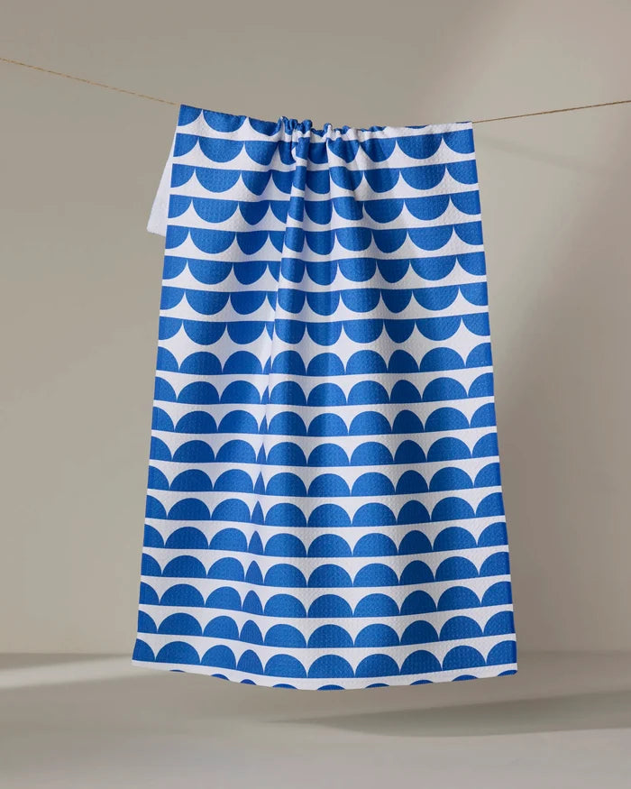 The Buy Guide's "Scallops - Navy" tea towel features a blue and white half-circle pattern and hangs against a plain, light backdrop.