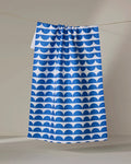 The Buy Guide's "Scallops - Navy" tea towel features a blue and white half-circle pattern and hangs against a plain, light backdrop.