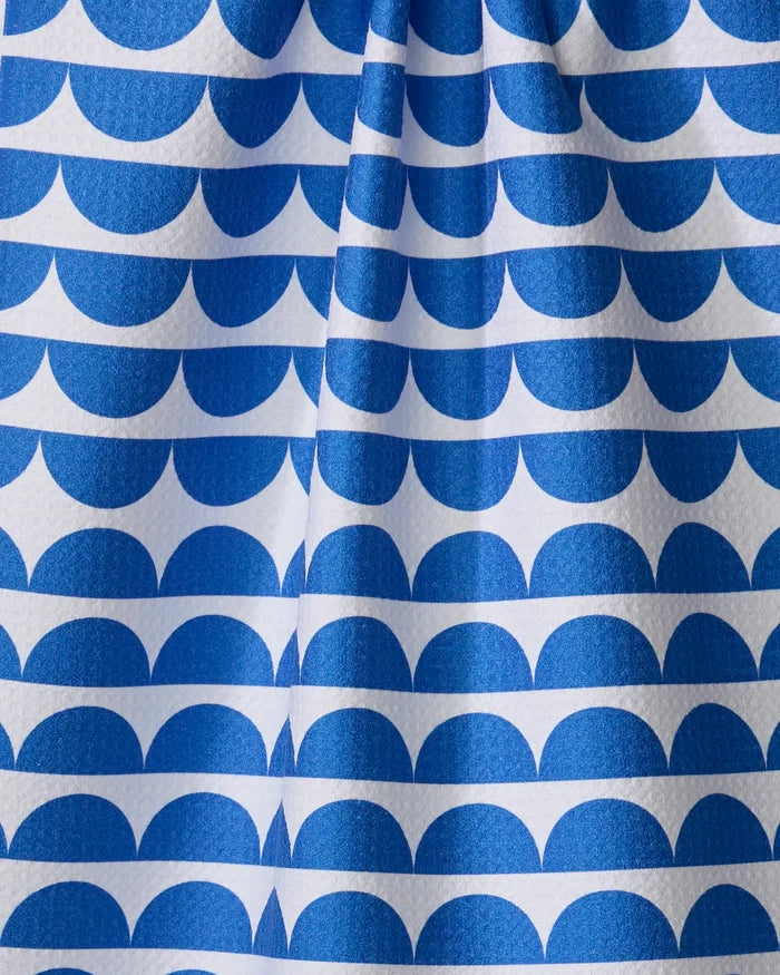 The Buy Guide's "Scallops - Navy" tea towel features a blue and white repeating half-circle wave pattern, with a central vertical fold that adds subtle texture and shadows.