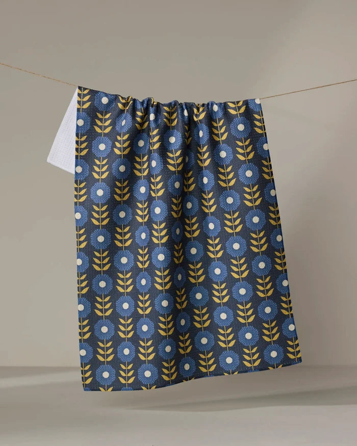 The Scandi Floral tea towel by Megan Kampa hangs on a clothesline, displaying a blue background with yellow leafy stems and white circular flowers in a repeating pattern.