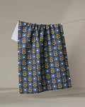 The Scandi Floral tea towel by Megan Kampa hangs on a clothesline, displaying a blue background with yellow leafy stems and white circular flowers in a repeating pattern.