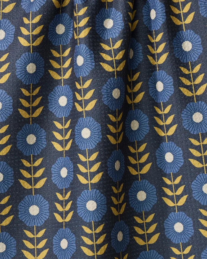 The Megan Kampa "Scandi Floral" kitchen towel features a pleated, eco-friendly fabric with a dark background and a repeating blue flower pattern with white centers and yellow stems for style and function.